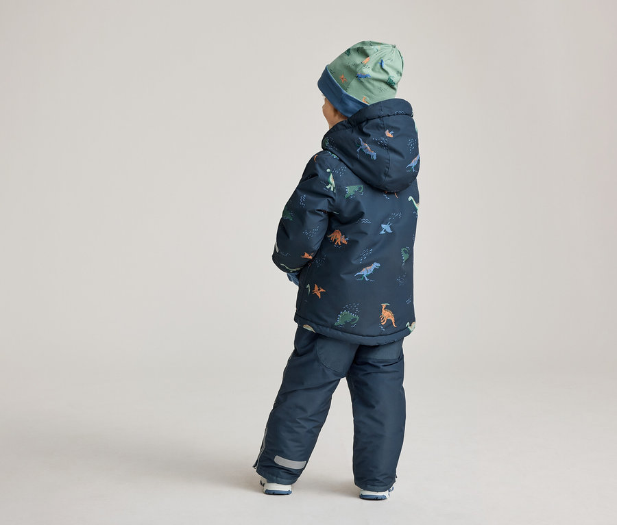 Un enfant, vu de dos, porte un ensemble bleu marine à motifs de dinosaures et un bonnet vert.
