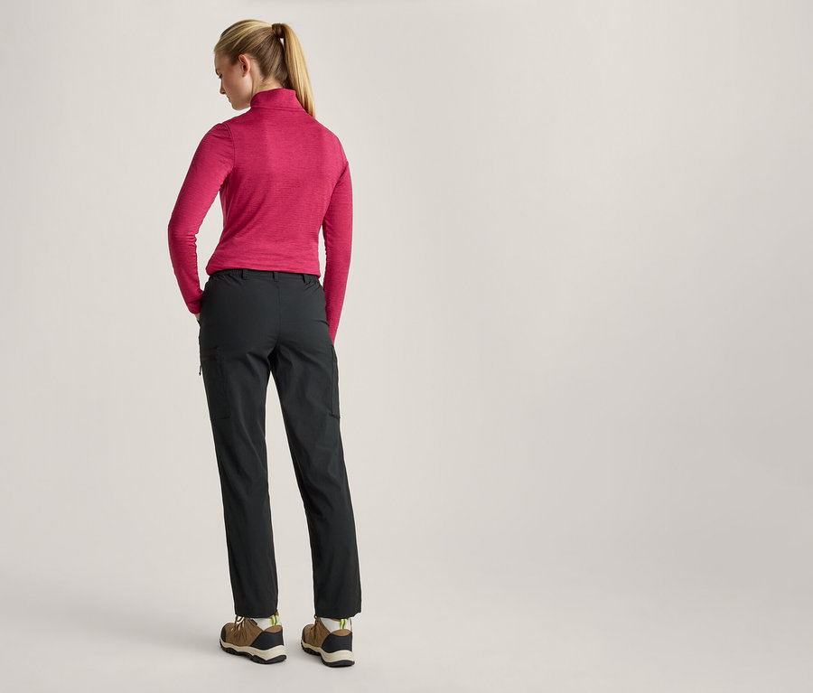 Femme de dos, portant un haut rose à manches longues, un pantalon gris foncé et des chaussures de randonnée, les mains dans les poches.