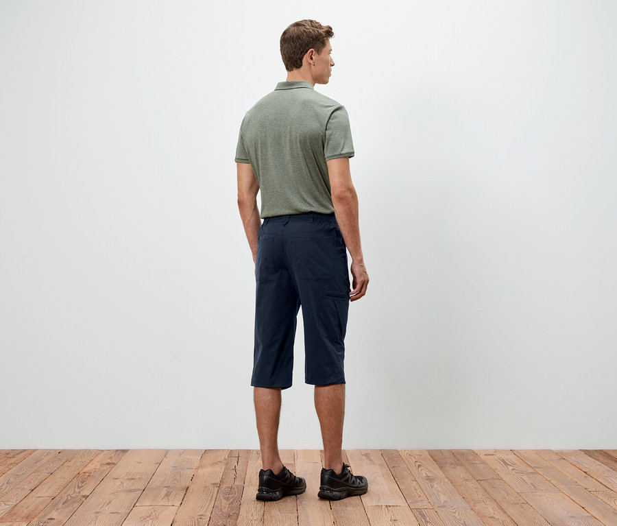 Un homme se tient dos à la caméra, portant un polo vert, un short bleu et des chaussures noires.