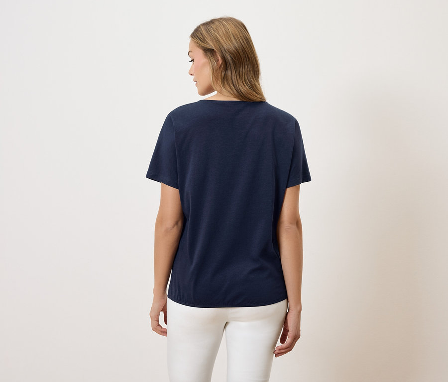Une femme aux cheveux blonds porte un t-shirt bleu marine et un pantalon blanc.