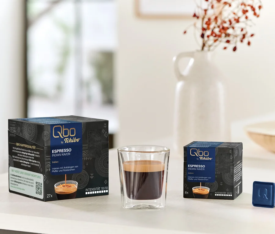 Sur une table, une tasse d'espresso et des paquets de Qbo Espresso INDIAN KAVERI avec 27 et 8 capsules.