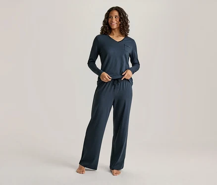 Une femme souriante aux cheveux foncés et bouclés porte un haut à manches longues et col en V bleu foncé avec une poche et un pantalon large assorti avec un cordon de serrage. Ses mains sont près de sa taille, et elle est pieds nus.