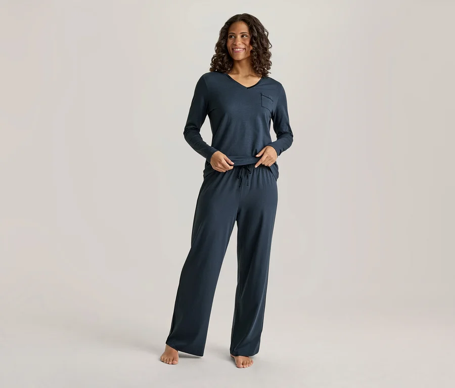 Une femme souriante aux cheveux foncés et bouclés porte un haut à manches longues et col en V bleu foncé avec une poche et un pantalon large assorti avec un cordon de serrage. Ses mains sont près de sa taille, et elle est pieds nus.