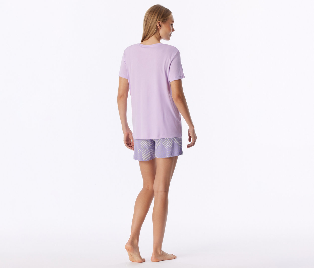 Femme debout, de dos, portant un t-shirt violet clair et un short à motifs géométriques clairs. Elle est pieds nus.