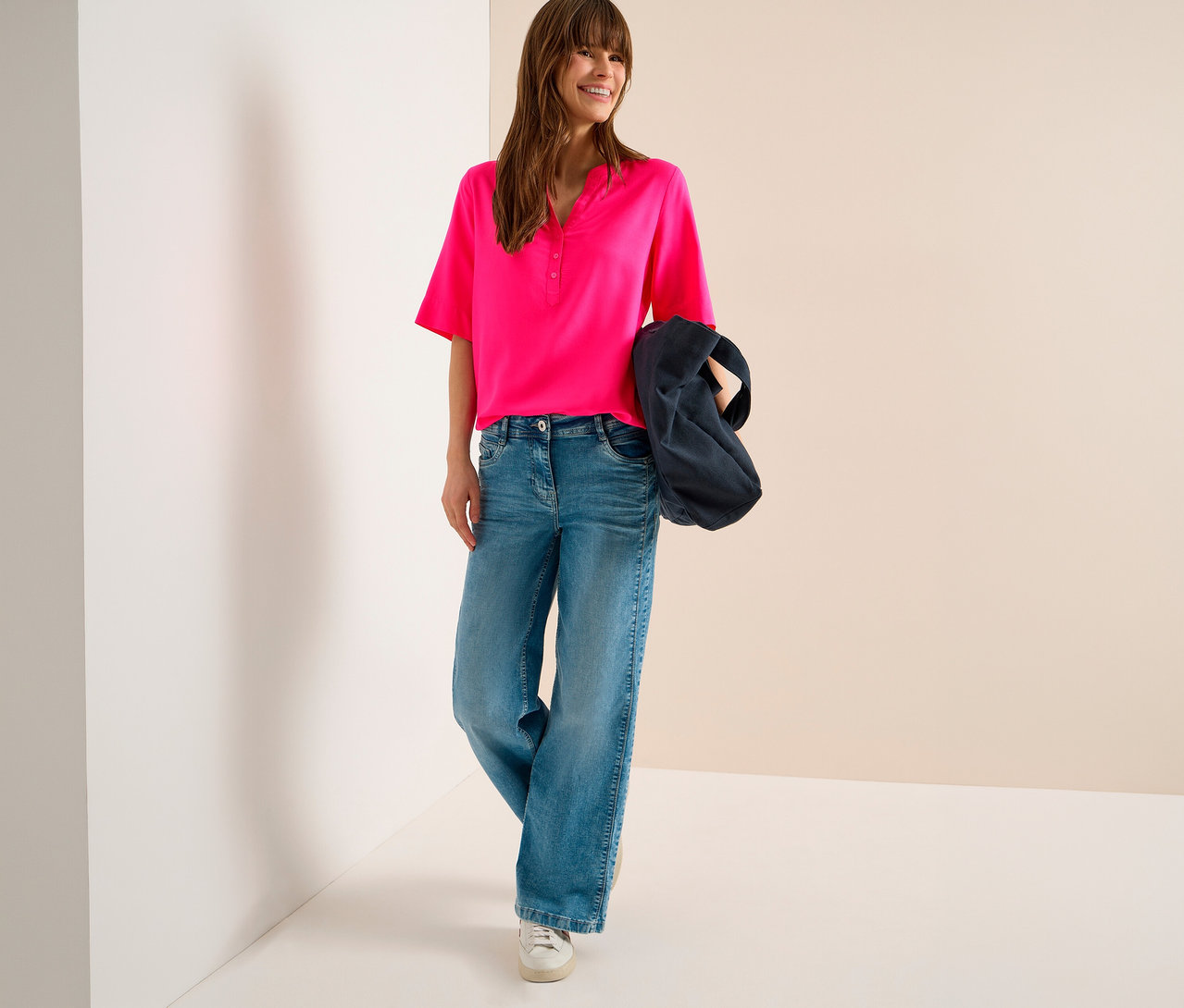 Femme souriante portant un chemisier tunique CECIL rose vif, un jean large bleu et des baskets blanches, tenant un sac foncé sur son bras.