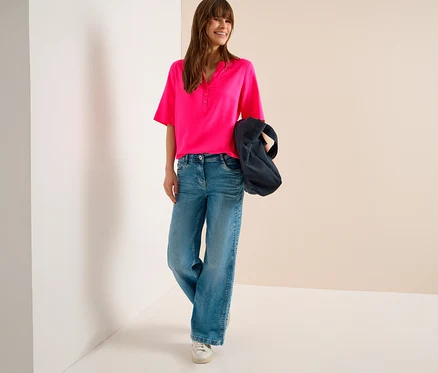 Femme souriante portant un chemisier tunique CECIL rose vif, un jean large bleu et des baskets blanches, tenant un sac foncé sur son bras.