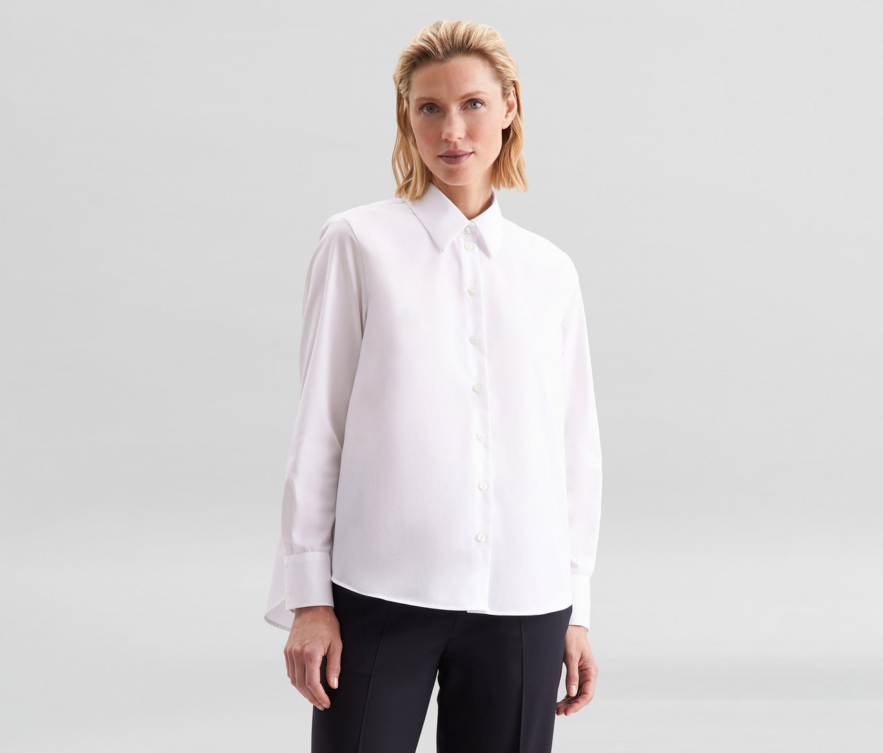 Femme aux cheveux blonds portant un chemisier sans repassage Seidensticker blanc et un pantalon noir, regardant devant elle.