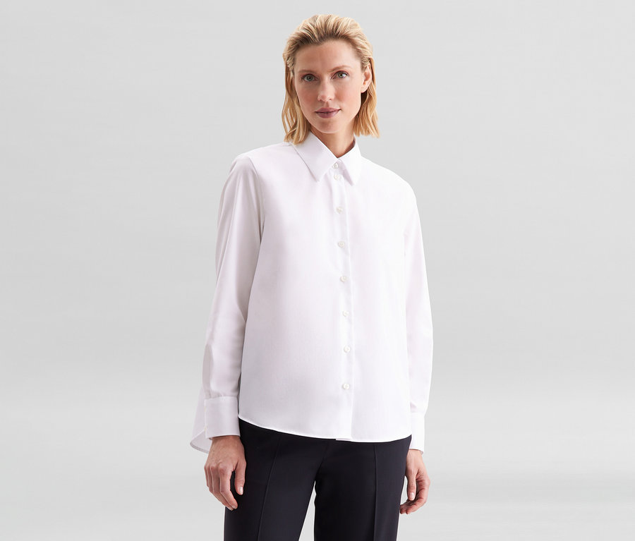 Femme aux cheveux blonds portant un chemisier sans repassage Seidensticker blanc et un pantalon noir, regardant devant elle.