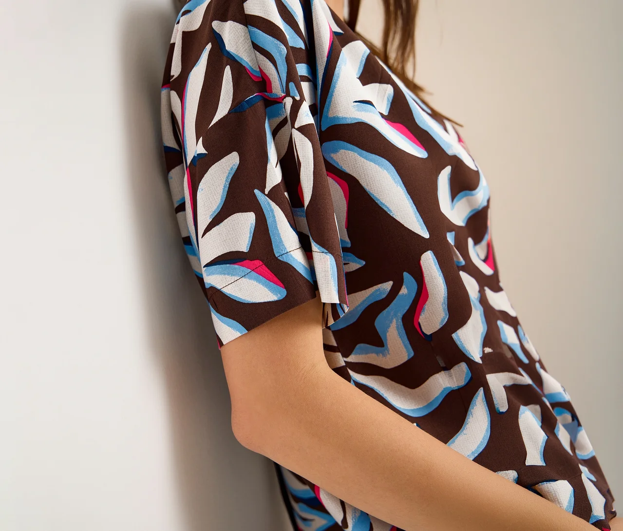 Une personne porte un chemisier à manches courtes CECIL à motif abstrait marron, blanc, bleu clair et rose, appuyée contre un mur blanc.