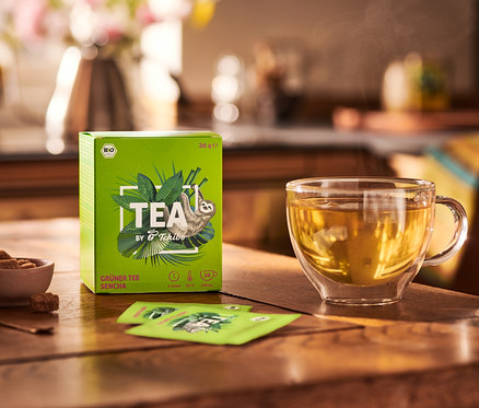Sur une table en bois se trouvent un paquet de BIO TEA by Tchibo Sencha 8x20x1,8g, un bol de sucre et une tasse de thé.