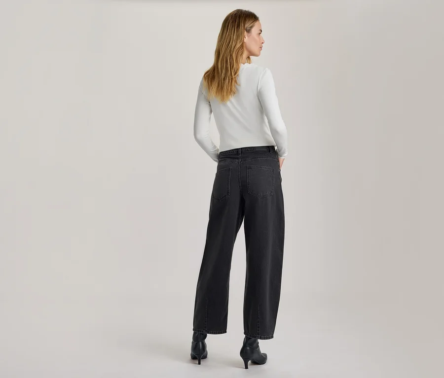 Femme vue de dos, légèrement tournée, portant un haut blanc à manches longues, un jean large gris foncé et des bottines noires à talons. Ses mains sont dans ses poches.