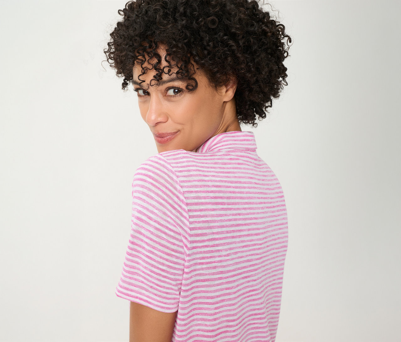 Femme aux cheveux bouclés, regardant par-dessus son épaule, portant un Polo Olsen rayé rose et blanc.