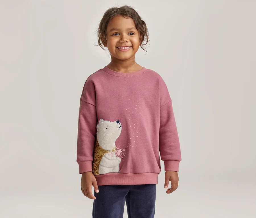Un jeune enfant souriant aux cheveux bouclés foncés porte un sweat-shirt rose orné d'un hérisson blanc au corps doré et texturé, regardant des paillettes blanches. L'enfant porte également un pantalon bleu foncé.