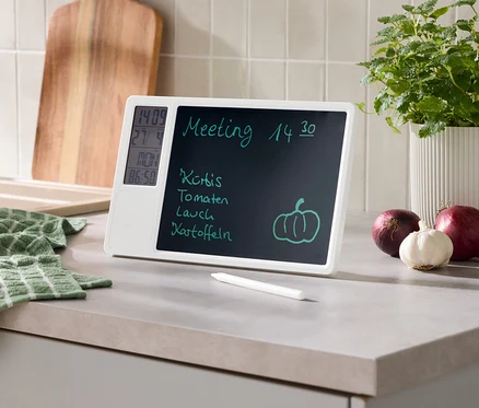 Un tableau mémo numérique blanc sur un comptoir de cuisine affiche une réunion, une liste de courses, l'heure, la date et la température. Un stylet, une serviette verte, une planche à découper en bois, une plante en pot, des oignons rouges et de l'ail sont à côté.