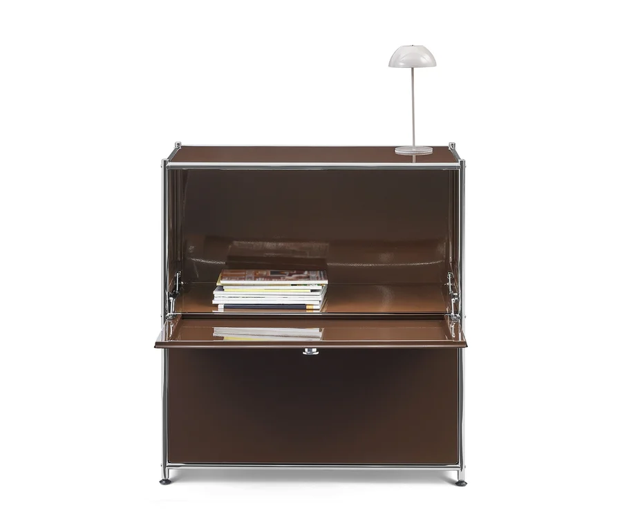 Secrétaire modulaire marron avec cadre chromé, sa porte abattante ouverte révélant une pile de magazines. Une petite lampe de table blanche est posée dessus.