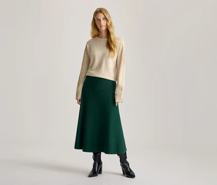 Femme aux longs cheveux blonds, portant un pull ras-du-cou beige, une jupe midi évasée vert foncé et des bottes noires à talons.