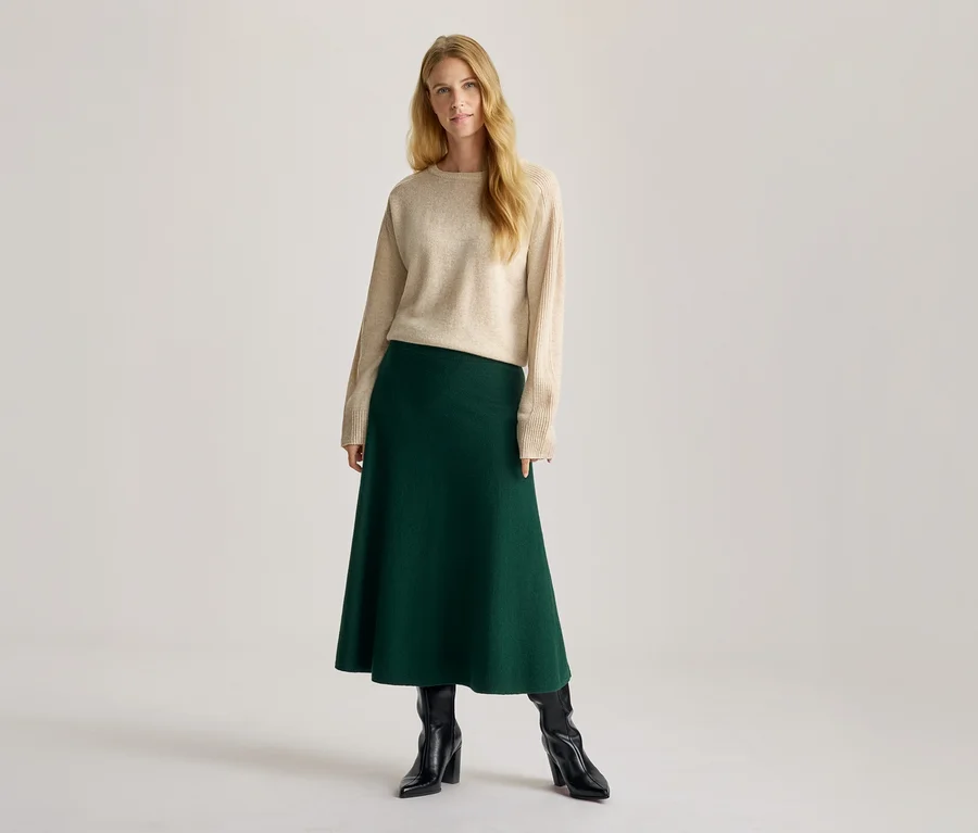 Femme aux longs cheveux blonds, portant un pull ras-du-cou beige, une jupe midi évasée vert foncé et des bottes noires à talons.