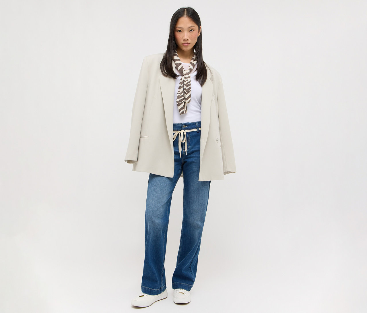 Jeune femme asiatique portant un t-shirt blanc, un jean large bleu avec un cordon de serrage clair, un blazer oversize beige sur les épaules et un foulard à motifs marron et blanc. Elle porte des chaussures plates blanches.