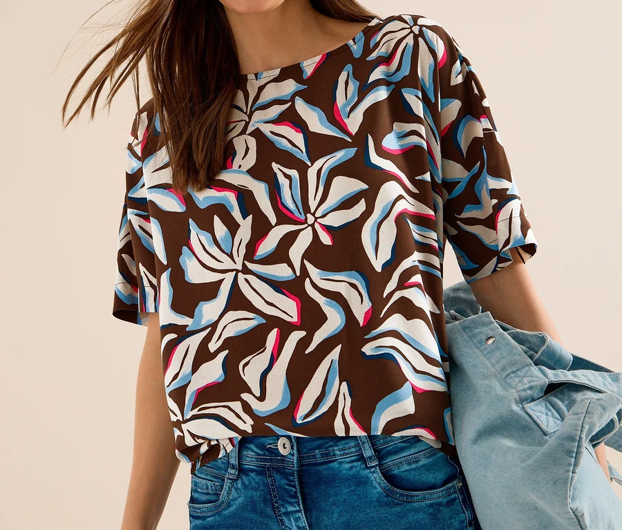 Femme souriante portant un chemisier à manches courtes CECIL marron à motifs floraux blancs, bleus et roses, un jean bleu et tenant une veste en jean.