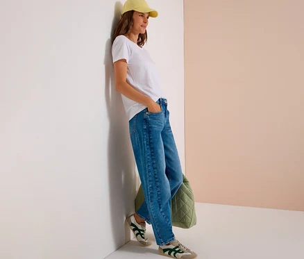 Femme appuyée contre un mur, portant un t-shirt blanc, une casquette jaune et un jean CECIL bleu, la main dans la poche. Un sac matelassé vert est au sol.