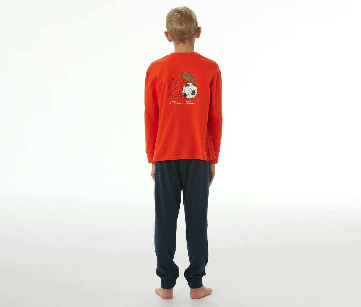 Un garçon est debout, dos à la caméra, portant un pyjama orange à manches longues et un pantalon de pyjama bleu marine. Le dos du haut est imprimé d'un ballon de basket, d'un ballon de football et d'un ballon de football américain, avec l'inscription « Power Team ».