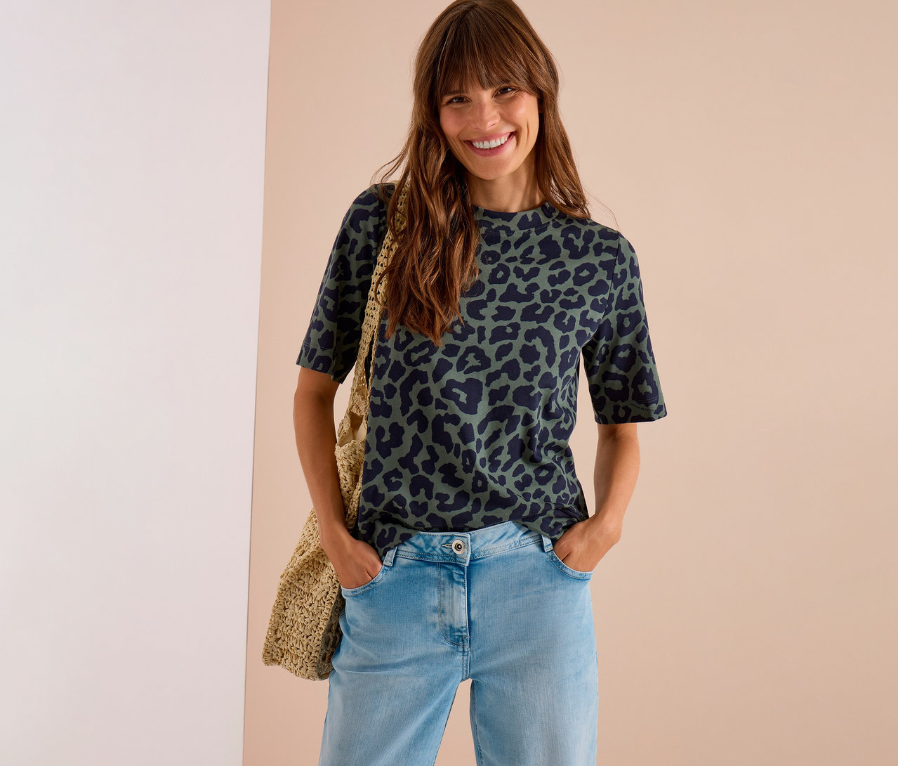Femme souriante aux cheveux longs portant un T-shirt en jersey CECIL vert foncé à imprimé léopard, un jean clair et un sac tressé.