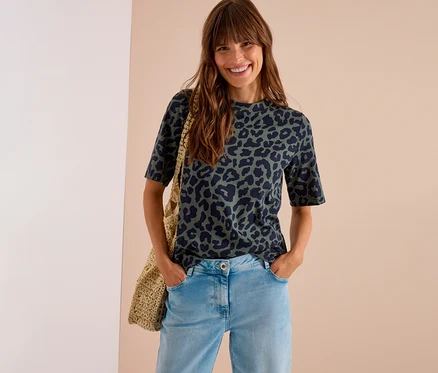 Femme souriante aux cheveux longs portant un T-shirt en jersey CECIL vert foncé à imprimé léopard, un jean clair et un sac tressé.
