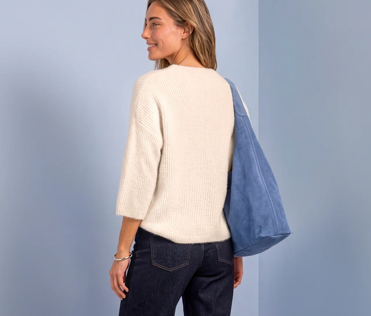 Femme de dos portant un pull en maille Street One crème, un jean foncé et un sac en daim bleu, regardant par-dessus son épaule.