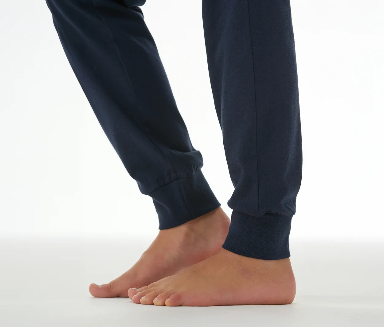 Bas des jambes et pieds nus d'une personne portant un pantalon de pyjama bleu foncé avec des poignets élastiques, debout sur une surface blanche.