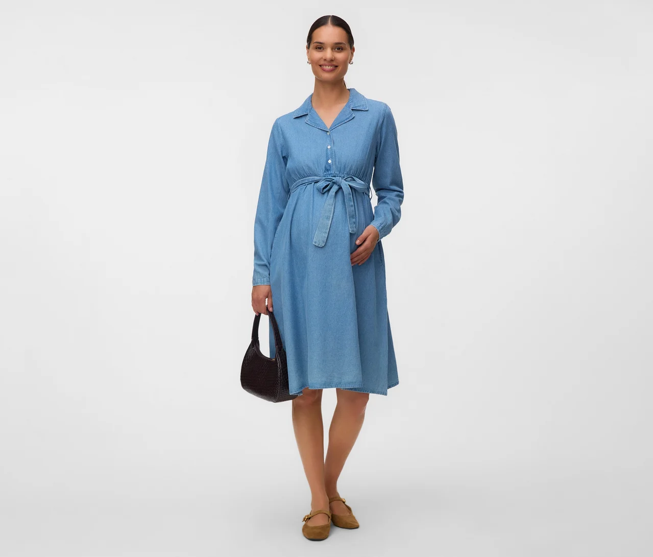 Femme enceinte portant une robe chemise en jean bleu avec une ceinture à nouer, un sac à main marron foncé et des chaussures plates marron clair.