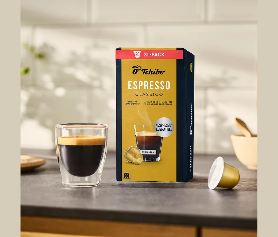Un paquet de 20 capsules Tchibo Espresso Classico est posé à côté d'un verre d'espresso avec une crème dorée et d'une capsule individuelle sur un comptoir de cuisine.
