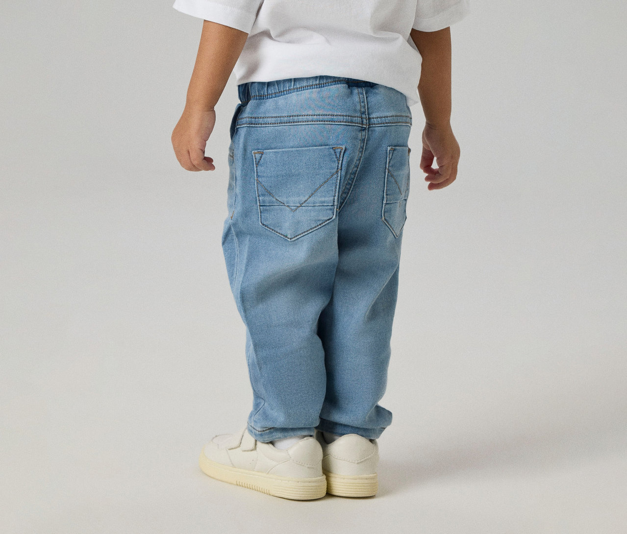 Enfant de dos portant un jean NAME IT Slim Sweat Jeans bleu clair avec poches arrière et des baskets blanches.