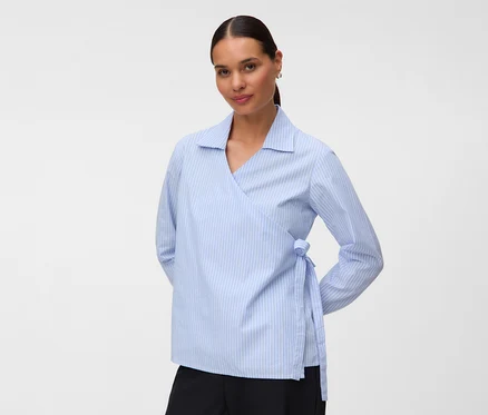 Femme aux cheveux foncés attachés en arrière, portant une blouse cache-cœur rayée bleu et blanc et un pantalon noir, regardant l'appareil photo.
