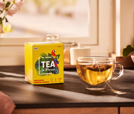 Paquet jaune de thé TEA by Tchibo infusion aux fruits bio curcuma-orange et une tasse de thé.