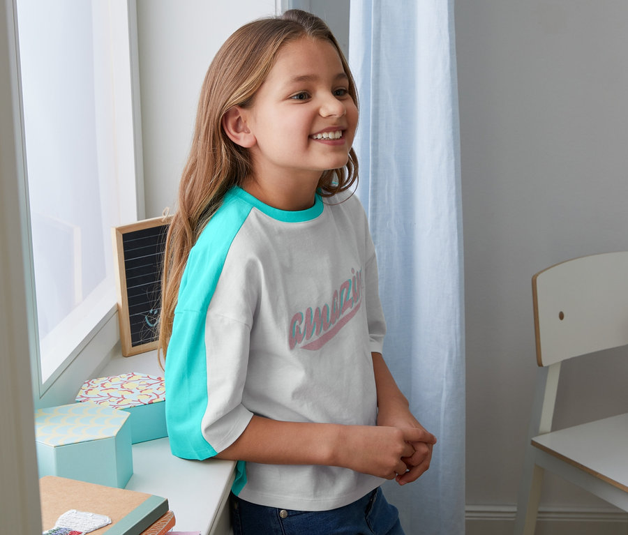 Une fille souriante s'appuie contre le rebord de la fenêtre et porte un t-shirt en coton bio avec des manches turquoise.