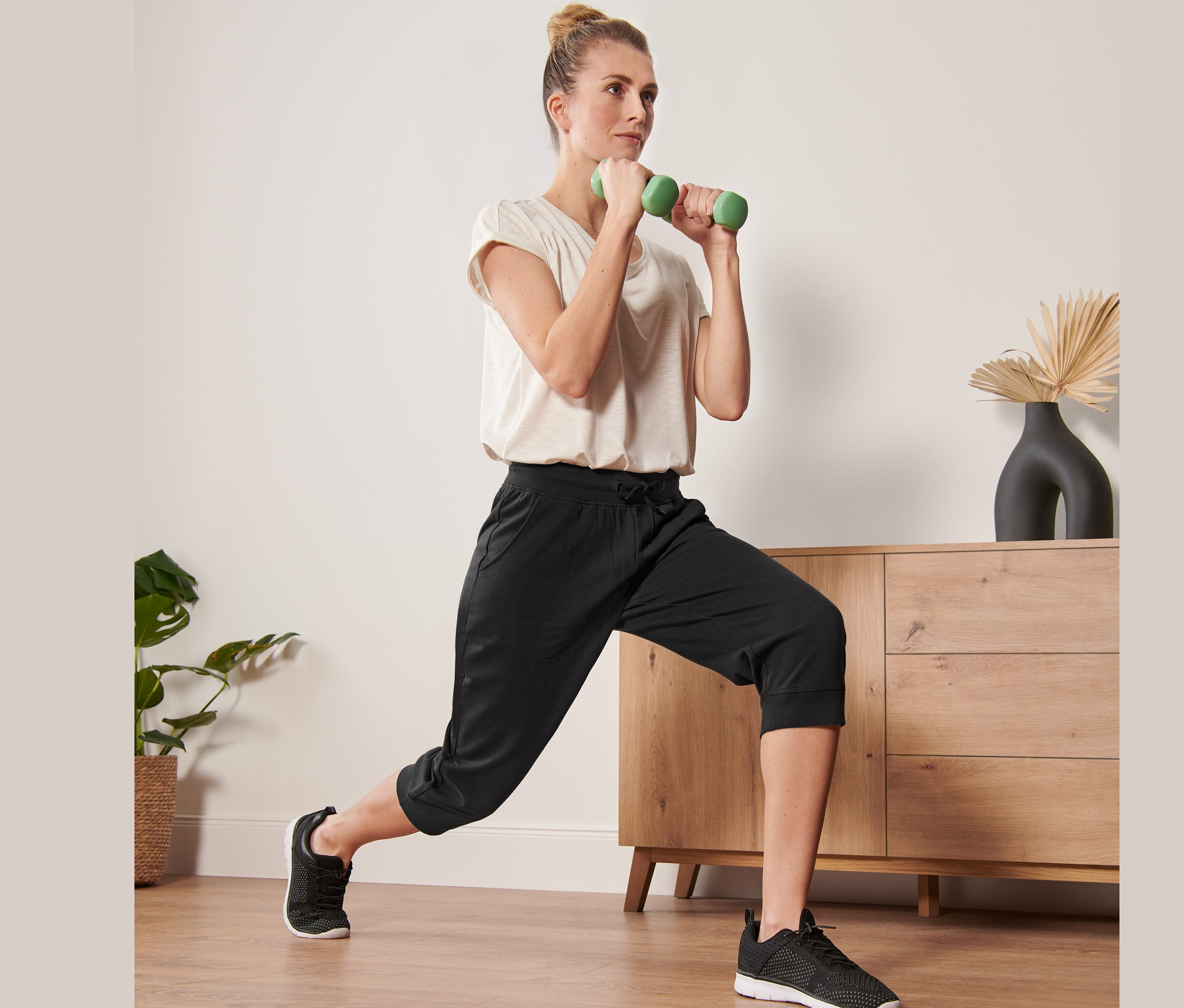 Femme faisant des fentes avec des haltères verts, portant un pantalon de sport 3/4 noir.