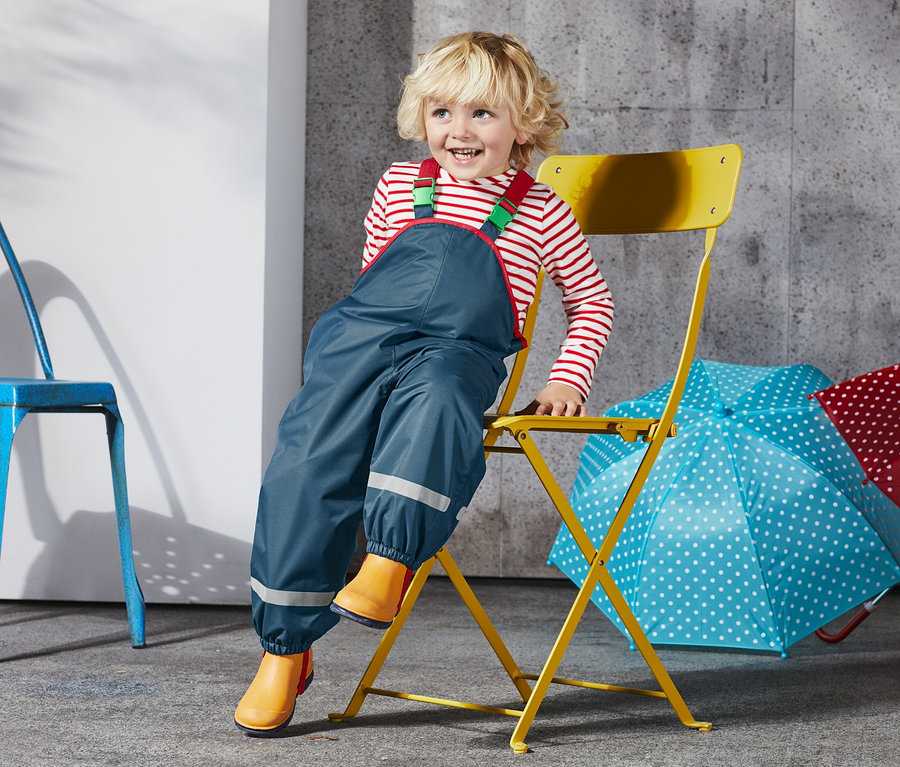 Un garçon est assis sur une chaise jaune et porte un pantalon de pluie bleu en matériaux recyclés.