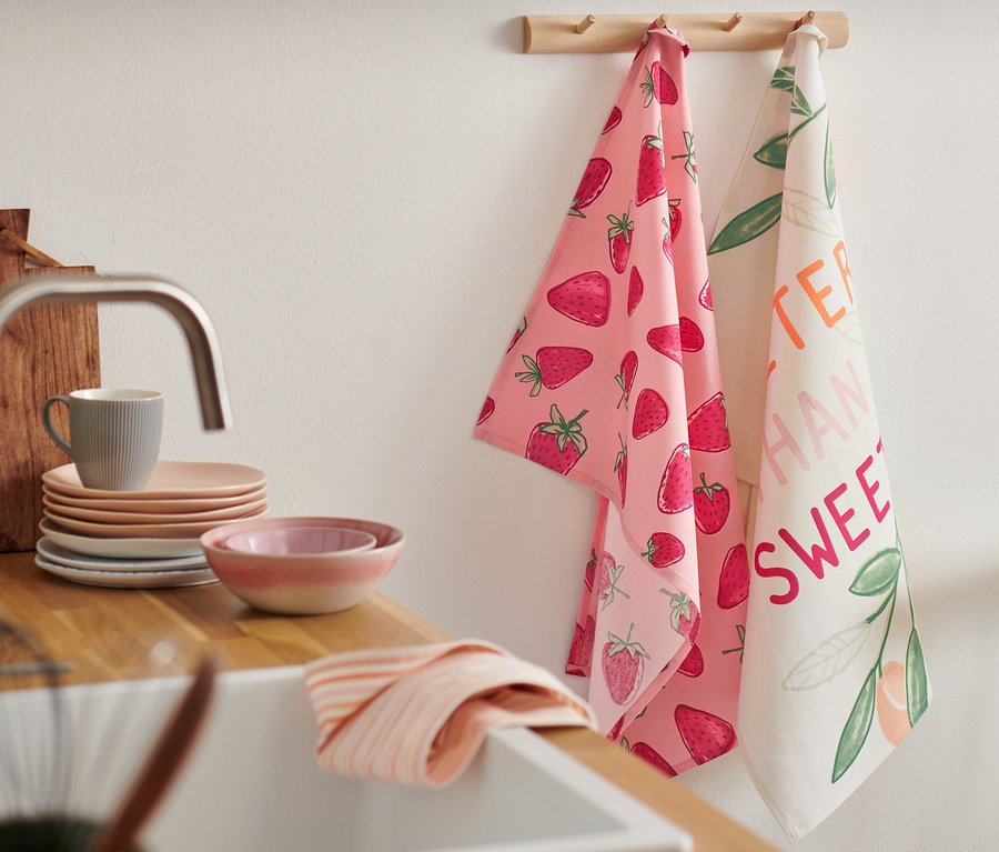 Deux linges de cuisine suspendus à une barre en bois. L'un est rose avec un motif de fraises, l'autre est blanc avec des inscriptions.