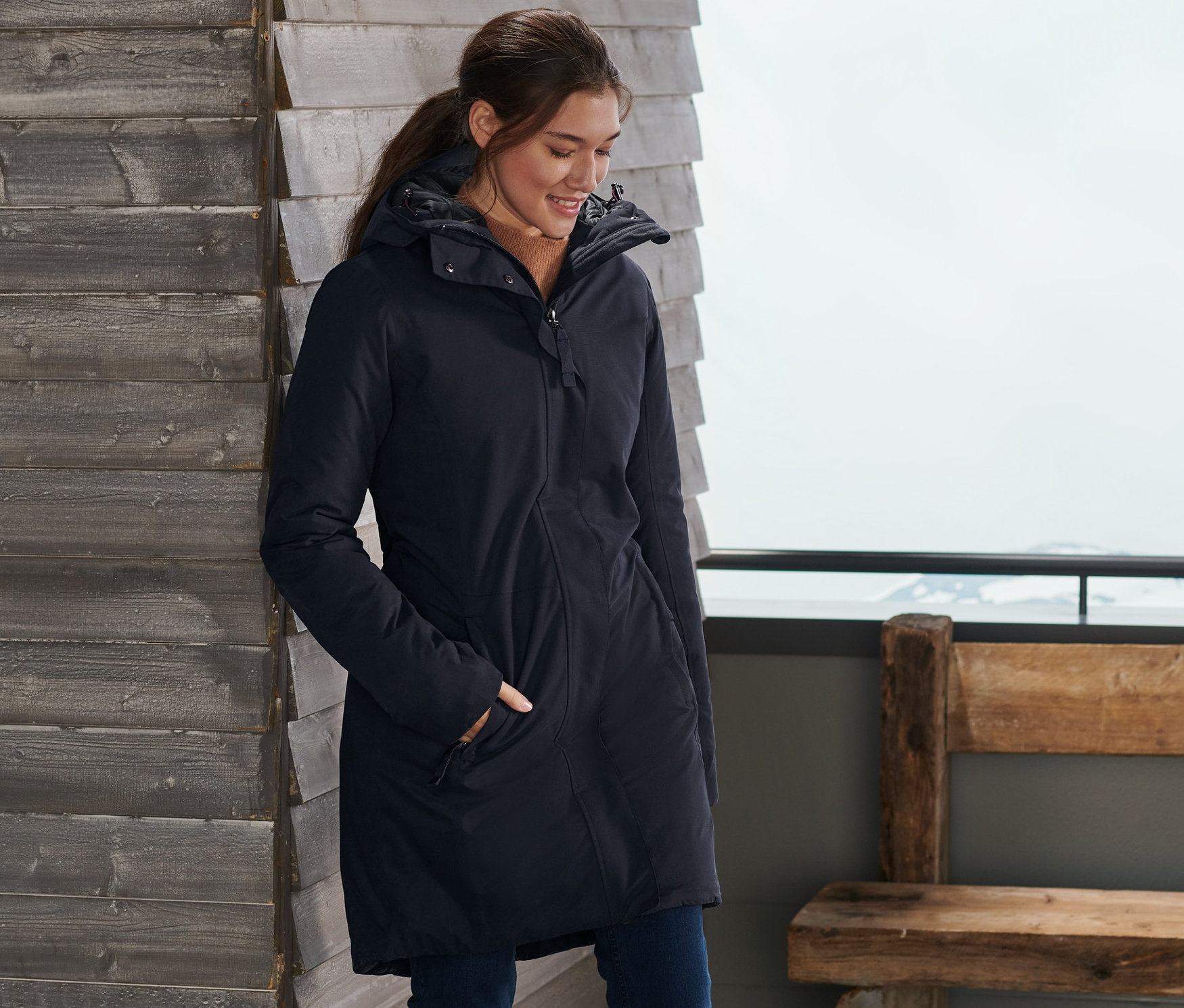 Une femme se tient près d'un mur en bois avec des skis et porte un manteau thermique bleu.