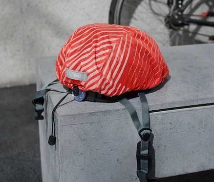 Une housse de pluie pour casque de vélo avec des rayures orange repose sur une surface en béton, avec un vélo en arrière-plan.