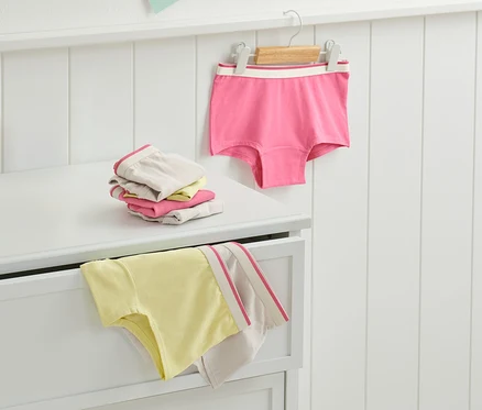 Sur une commode sont pliées 7 culottes roses pour filles, une autre est suspendue à un cintre.