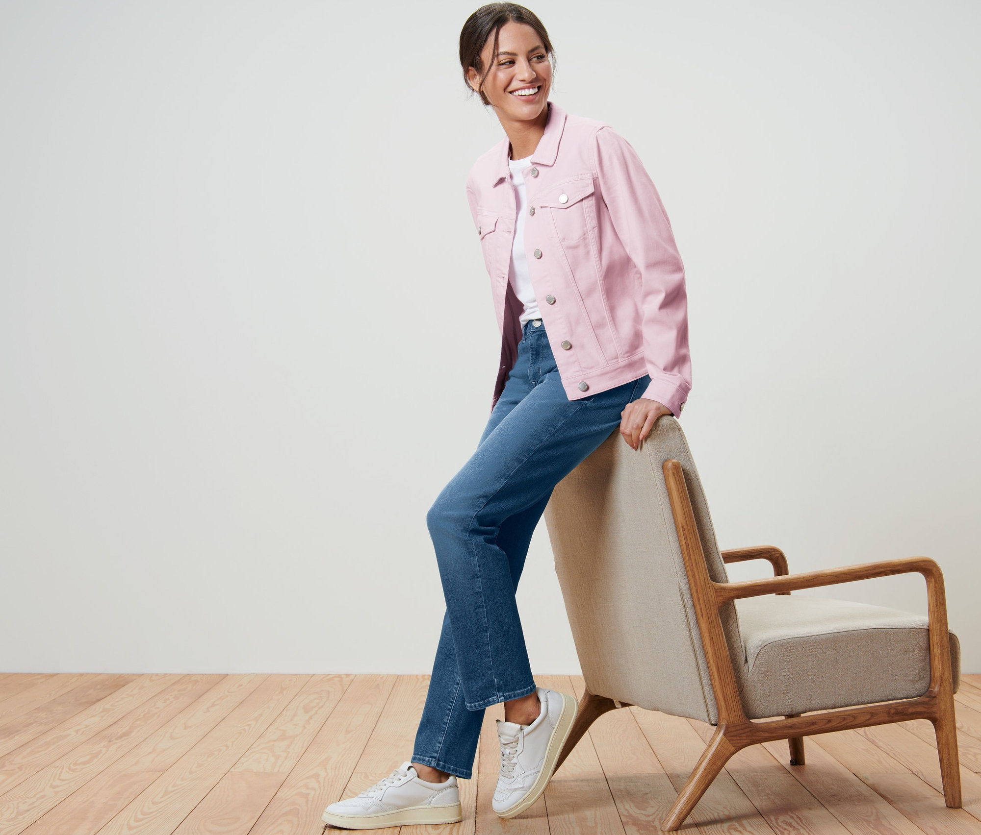 Femme souriante posant en jaquette en jeans rose et jeans à côté d'un fauteuil.