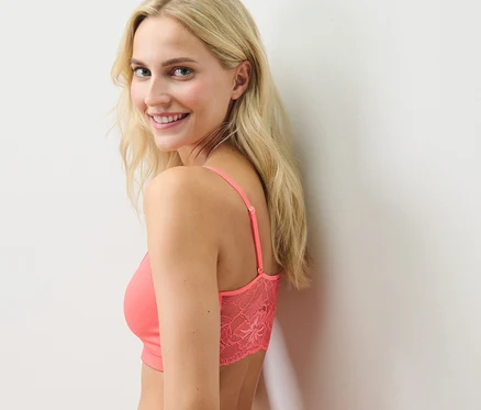 Une femme blonde souriant par-dessus son épaule porte une brassière seamless avec dentelle, couleur papaye.