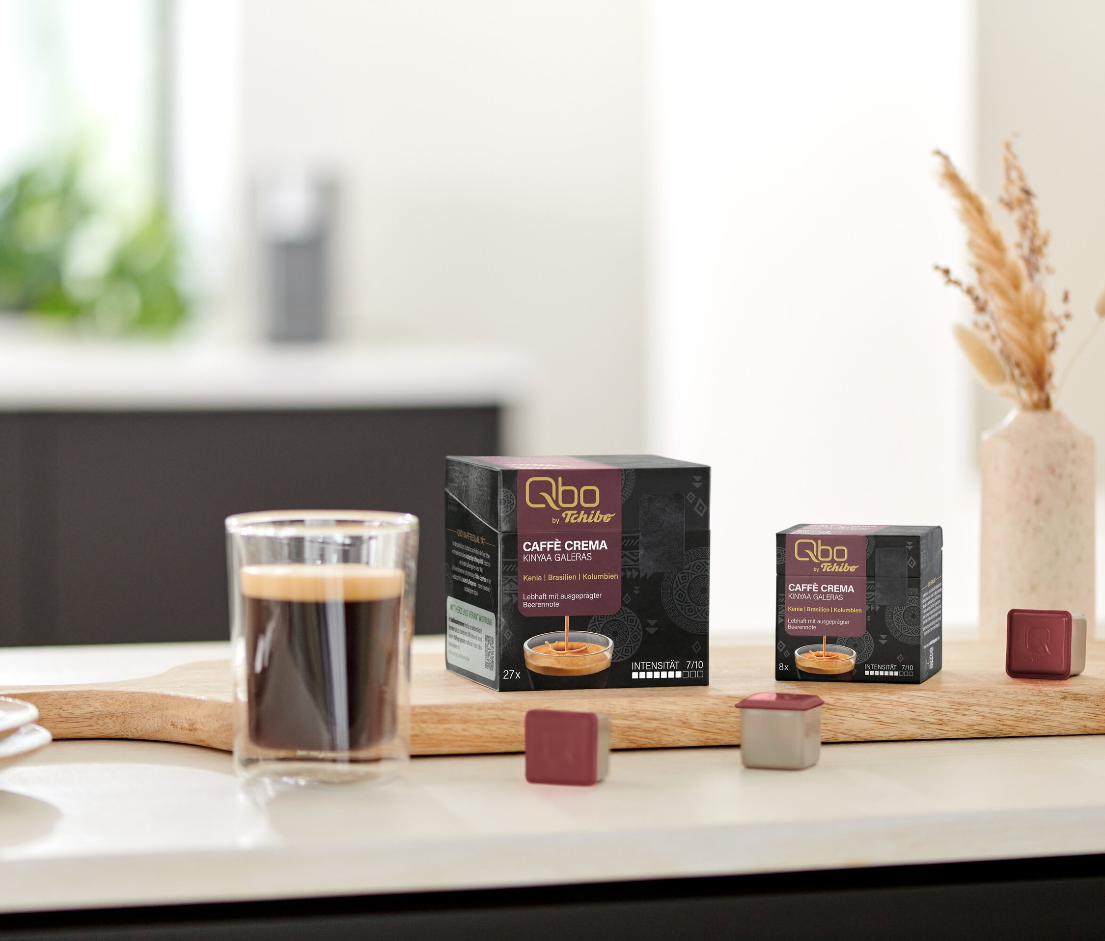 Un verre de café est posé à côté des paquets de capsules Qbo Caffè KINYAA GALERAS sur une planche en bois.