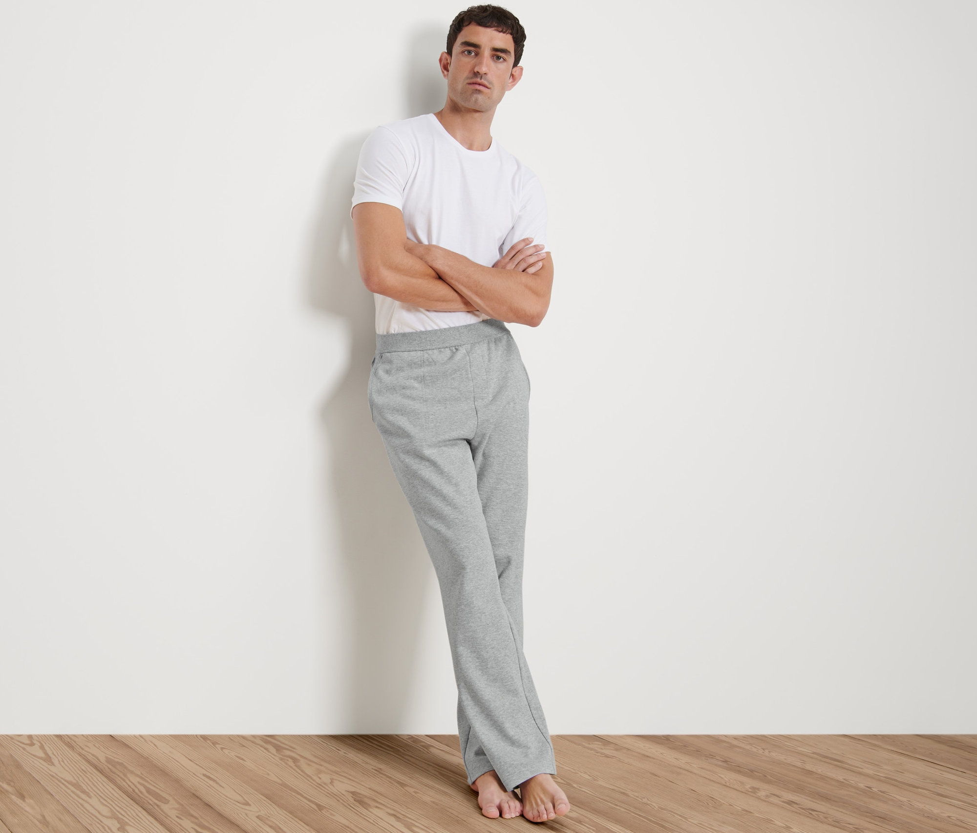 Un homme appuyé contre un mur porte un t-shirt blanc et un pantalon d’intérieur gris.