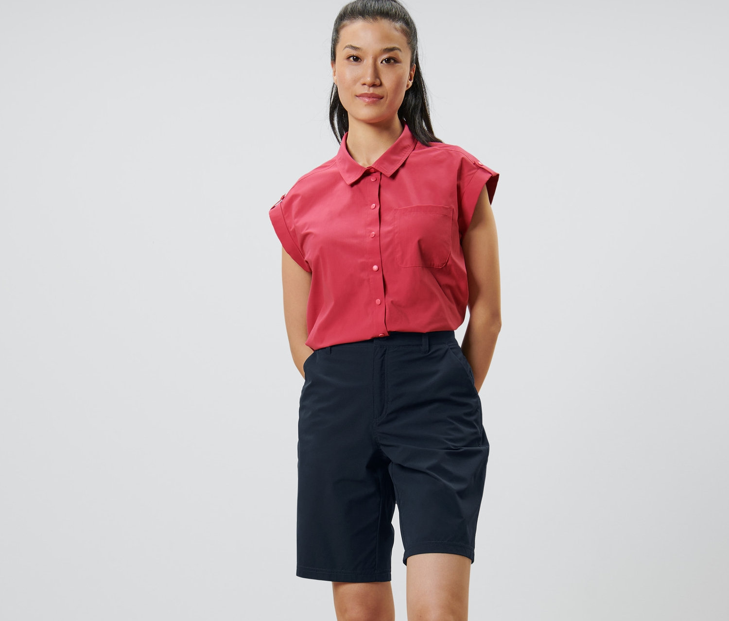 Une femme se tient debout, les mains derrière le dos. Elle porte un short fonctionnel en softshell bleu foncé et une chemise rouge.