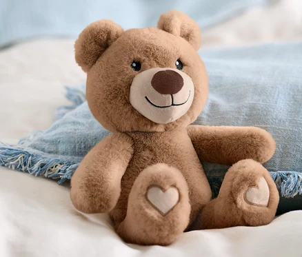Un animal en peluche « ours » marron est assis sur du linge de lit blanc et un coussin bleu.