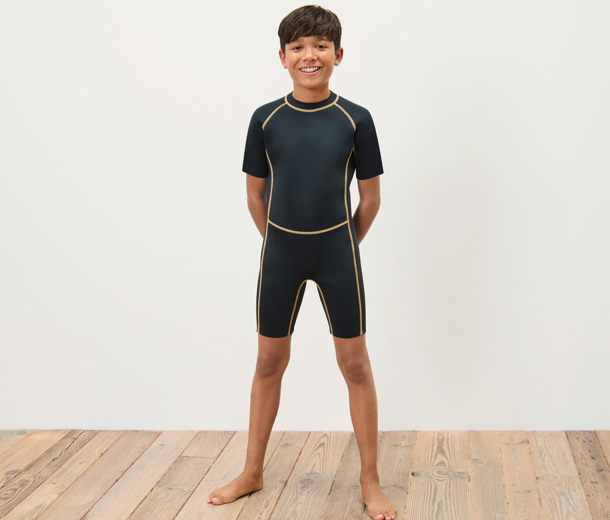 Un garçon en combinaison de néoprène pour enfant se tient pieds nus sur un plancher en bois devant un mur blanc.