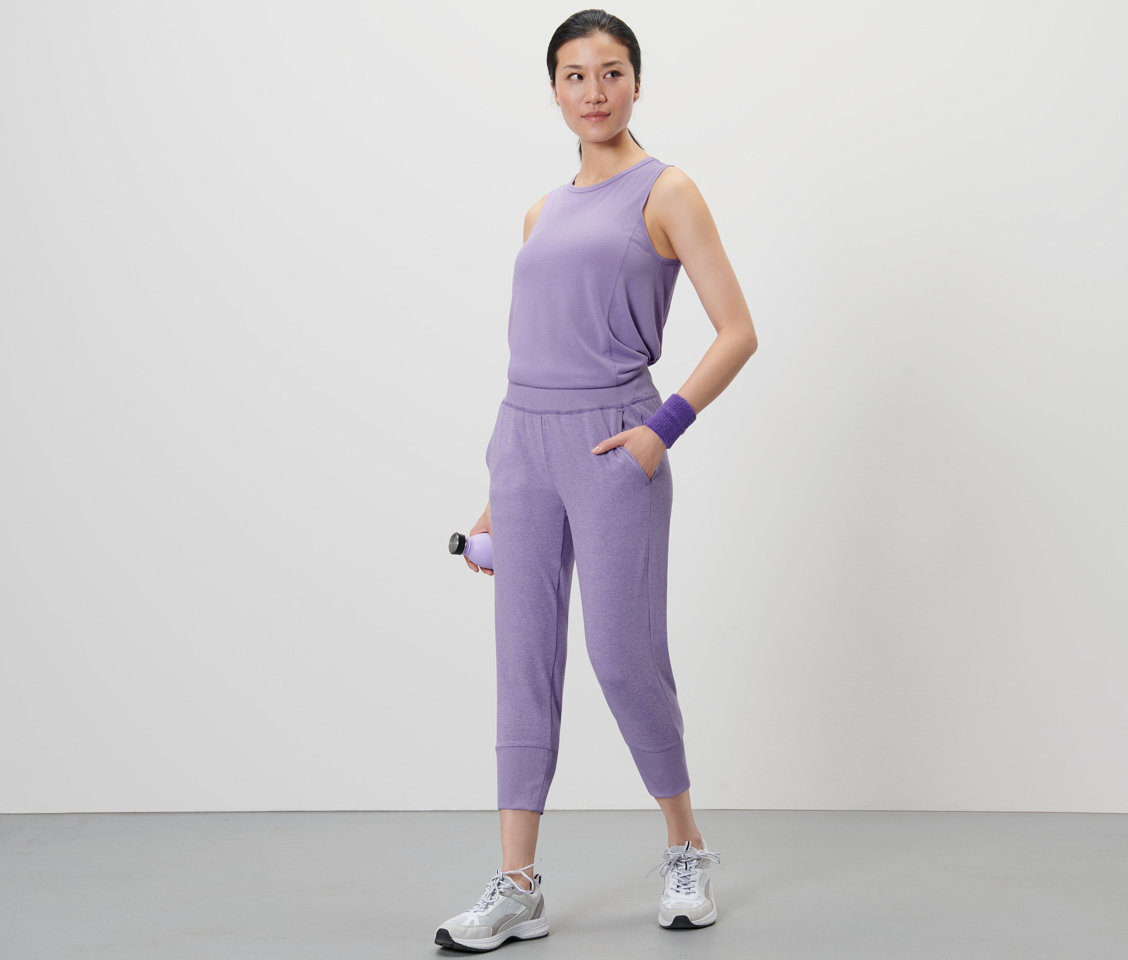 Une femme debout tenant une bouteille d'eau violette. Elle porte un haut violet et un pantalon de sport 3/4.
