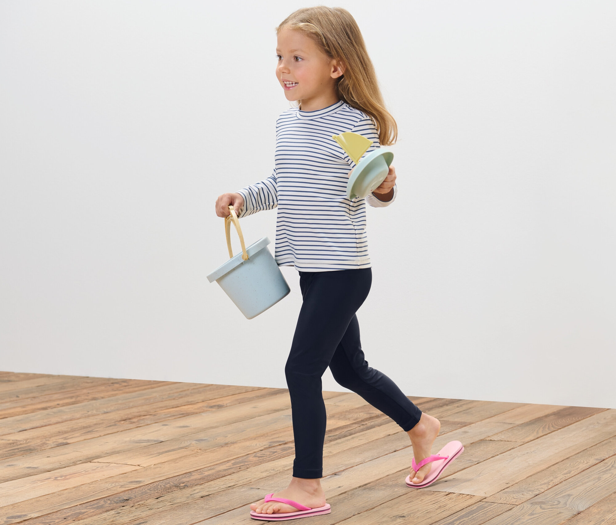 Une fille marche sur un plancher en bois et porte un seau bleu et des jouets de sable. Elle porte un legging de bain pour enfant avec un indice de protection UV 80.